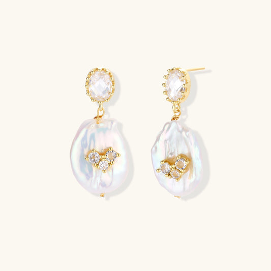 Gold-plated drop earrings with iridescent baroque pearls and crystal flower accents on cream background
